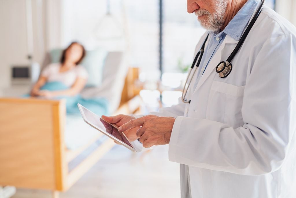 Photo of a doctor standing in hospital room, using tablet, with patient in a private hospital room. Concept of a safe detox from high-dose opioids.