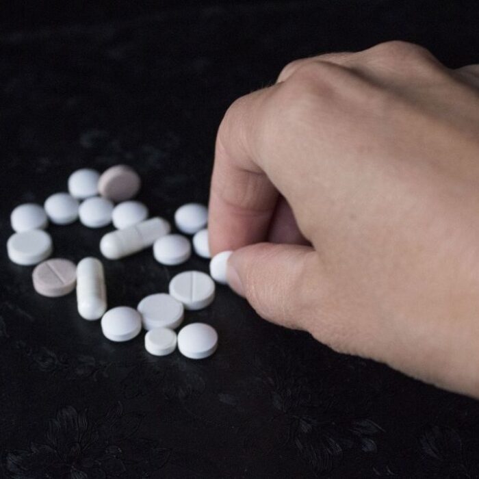 Scattered different pills and capsules close up on a black background with a hand picking up a pill. Concept of polydrug use.