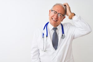 Senior grey-haired male doctor wearing stethoscope standing over isolated white background, uncertain with doubt, thinking with hand on head. Pain management doctor retiring concept.