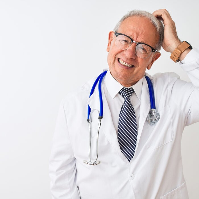 Senior grey-haired male doctor wearing stethoscope standing over isolated white background, uncertain with doubt, thinking with hand on head. Pain management doctor retiring concept.